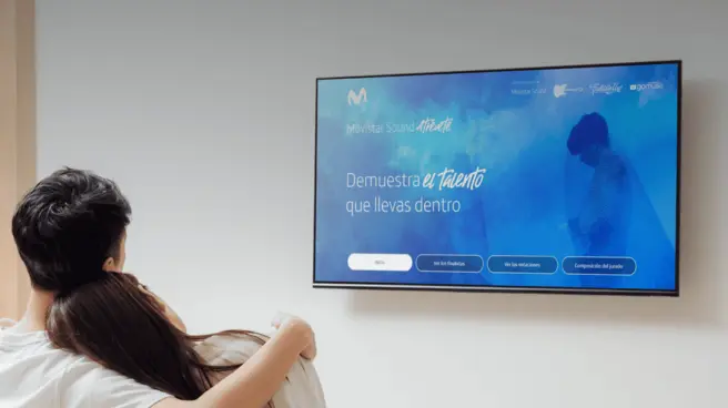 A couple sitting on a sofa watching the LivingApp Movistar platform on a television.